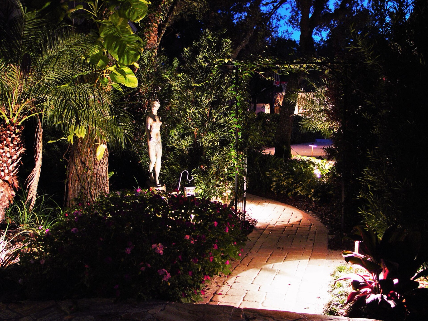 allée en briques qui tourne dans un jardin de nuit, éclairée au ras du sol par de petits points lumineux cachés dans les massifs fleuris, une statue éclairée à son pied